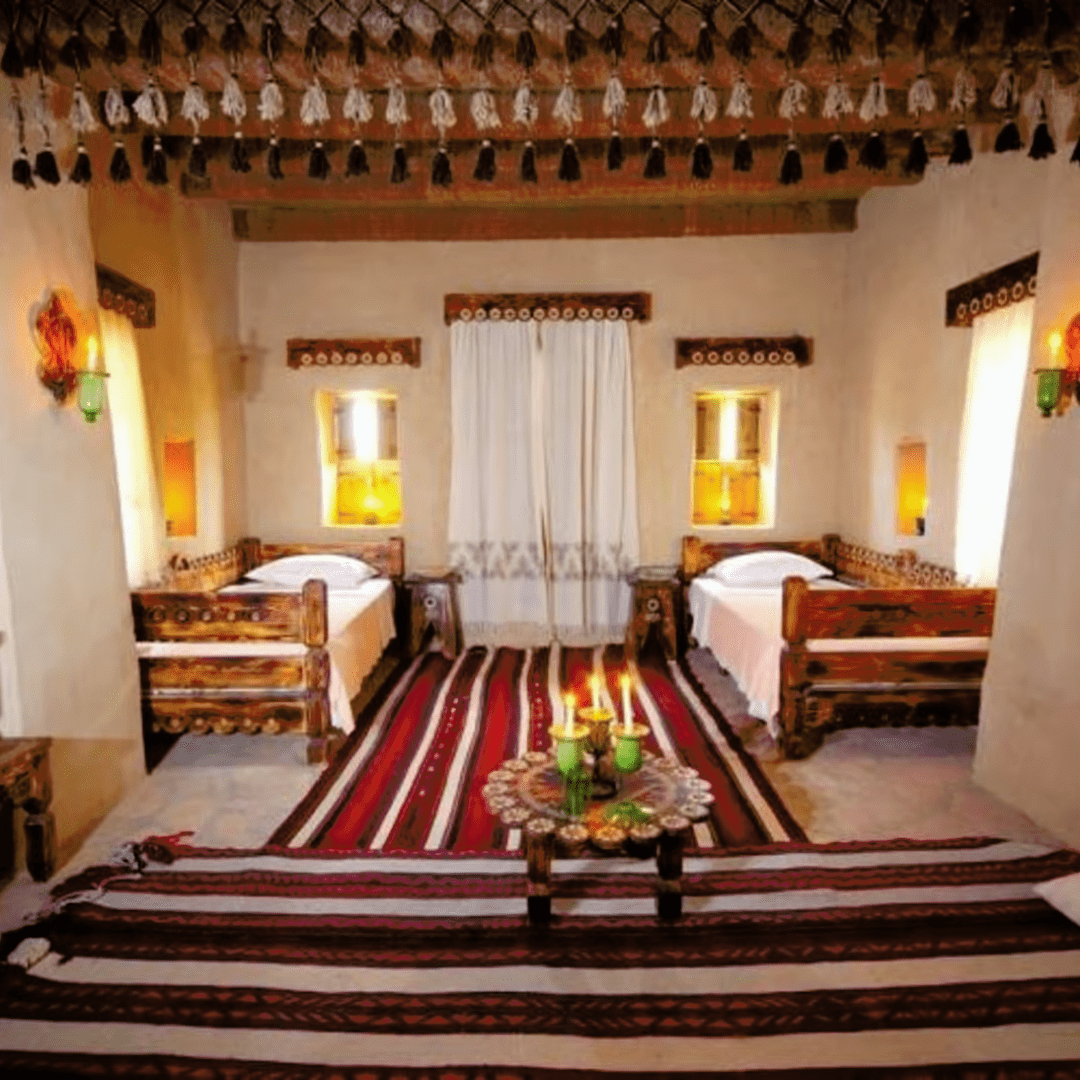 A cozy rustic bedroom with two single beds, each flanked by warm wall-lights, colorful striped rugs on the floor, a small table with lit candles in the center, and traditional decor elements.