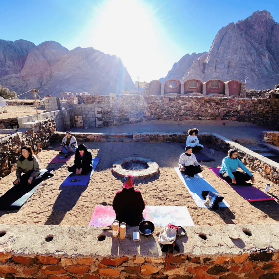 Outdoor yoga class with participants on mats in the egyptian desert setting with mountains in the background, under a clear sky with the sun shining.