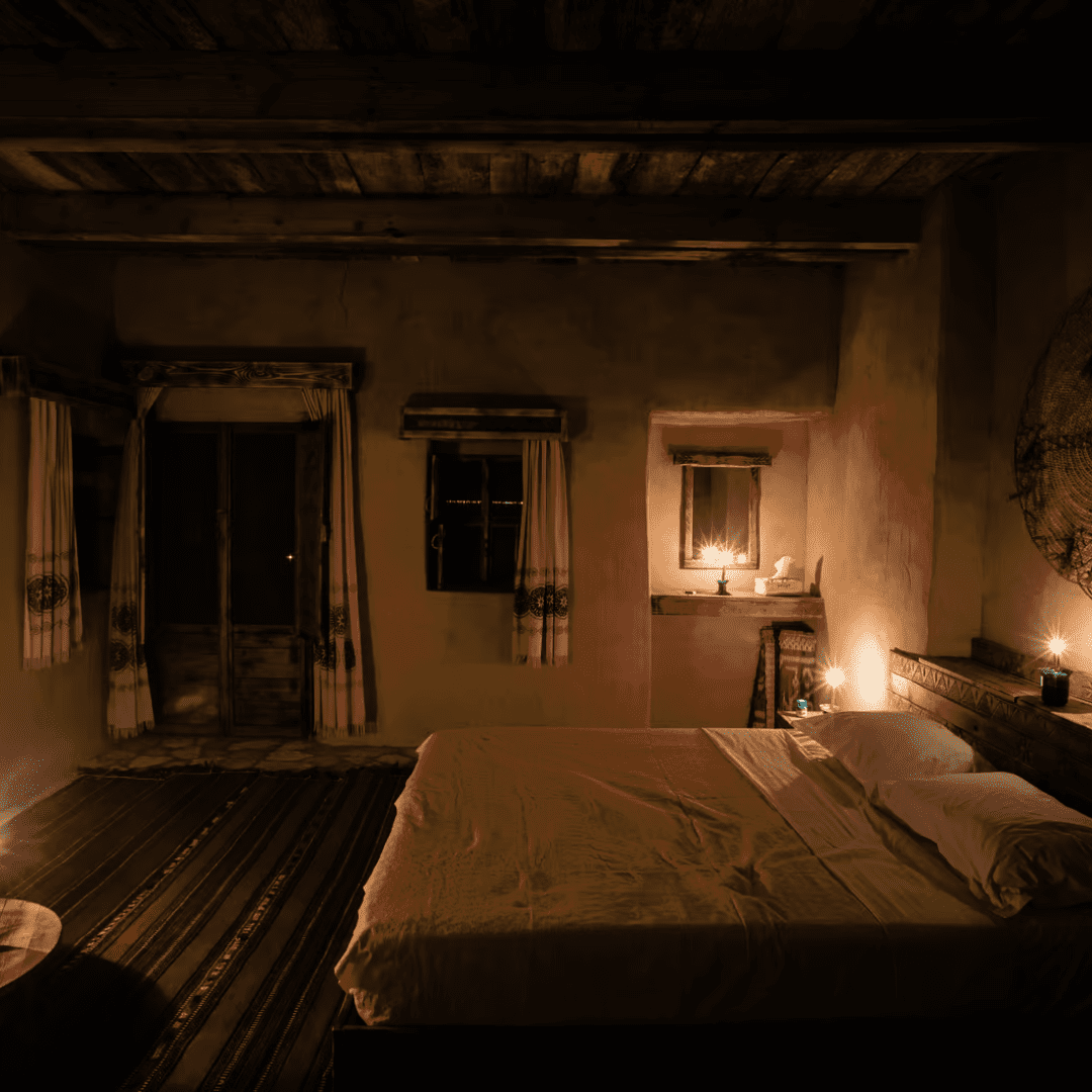 A dimly lit rustic bedroom with a large bed in the center, a patterned rug on the floor, decorated with multiple lit candles, and featuring a door and windows with sheer curtains.