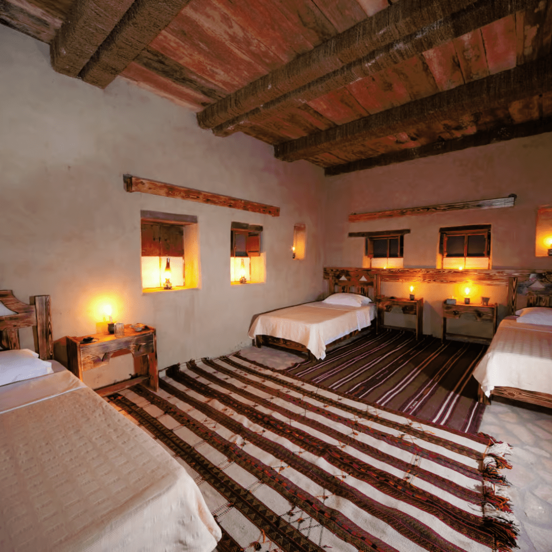 Alt text: A rustic bedroom with two single beds, wooden furniture, exposed wooden ceiling beams, and warm wall sconces casting a soft glow. The room is adorned with striped rugs on the floor and simple white bedding on the beds.