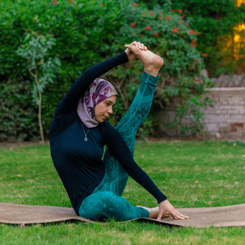 A woman wearing a hijab and exercise attire performs a seated yoga pose outdoors on a mat, with a natural green background.