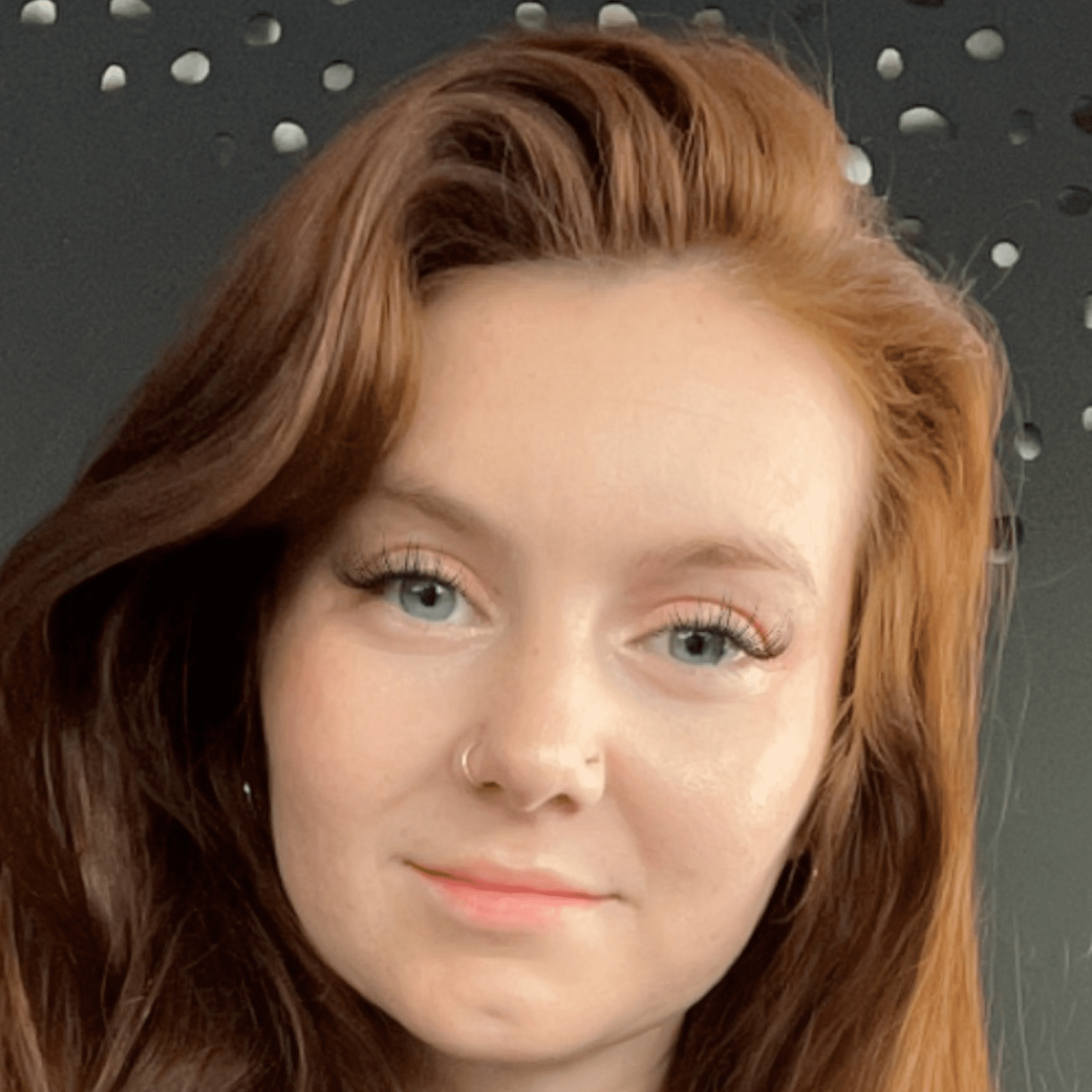A close-up photo of a smiling woman with long auburn hair, clear skin, a nose ring, and makeup showcasing defined eyelashes. She is in front of a dark background with a pattern of light circular shapes.