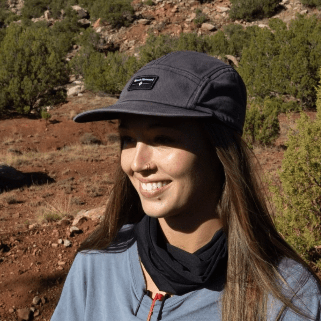A smiling woman wearing a black cap outdoors with a sunlit, rocky landscape and greenery in the background.